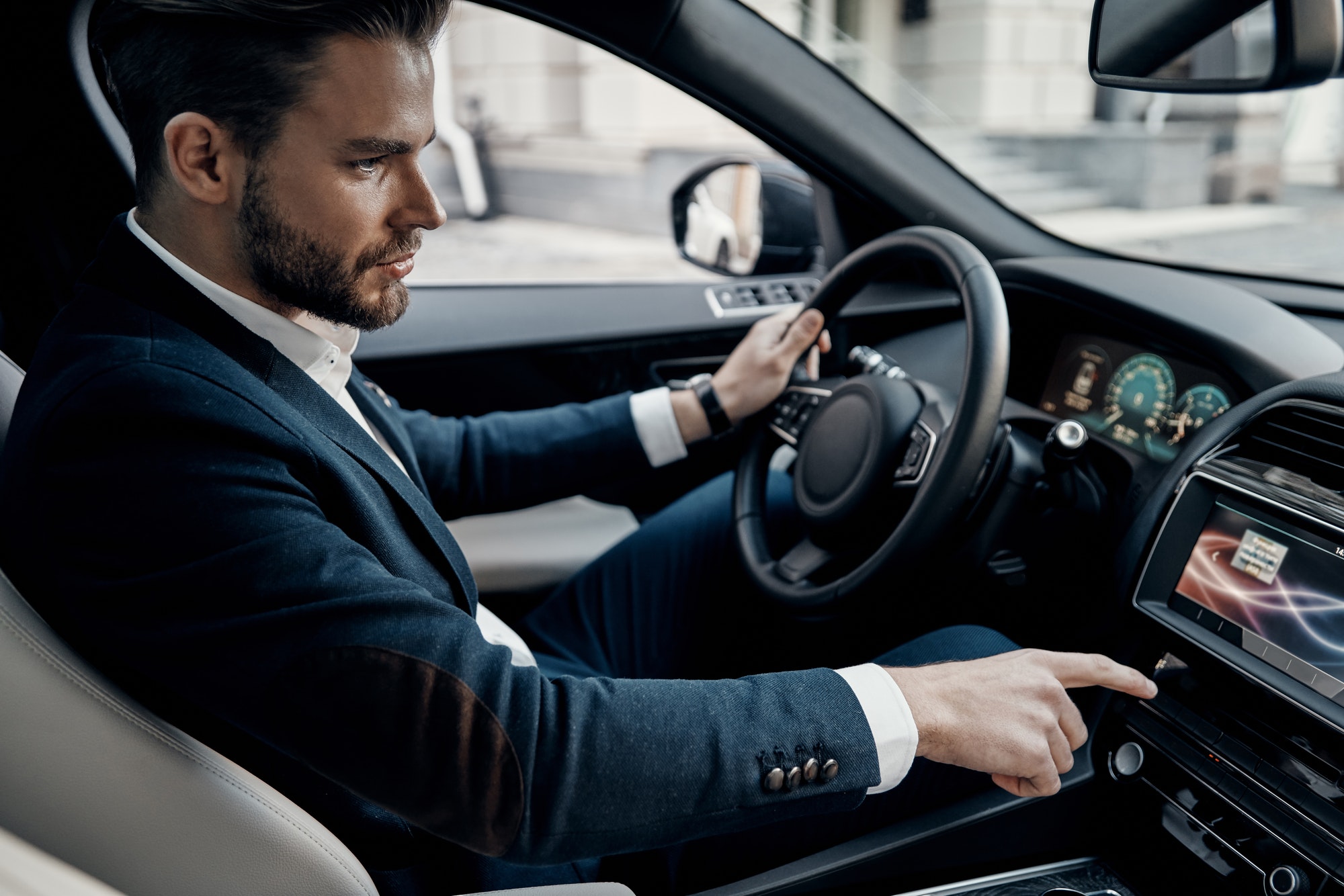 On the way to business meeting. Handsome young man in full suit pushing buttons while driving a car